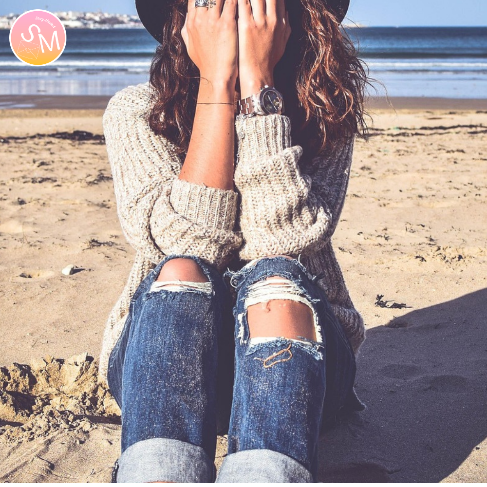 Person sitting on a beach wearing a sweater and ripped jeans, with a scenic background. heartbreak story delivered to your mailbox monthly